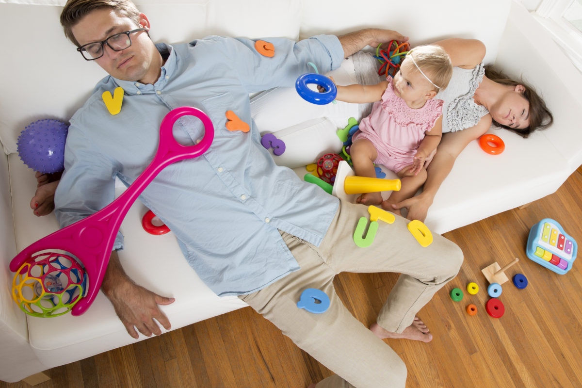 Parents sleeping on sofa while baby girl plays with toys